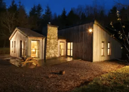 A modern cabin with wooden cladding and a stone chimney is warmly lit at night, surrounded by trees and an earth yard, creating a cosy and inviting atmosphere.