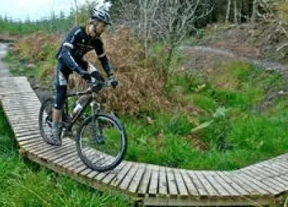 A cyclist wearing a helmet and cycling gear rides a mountain bike along a narrow, curved wooden trail through grassy and wooded terrain.