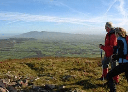 Two hikers with rucksacks and walking sticks stand on a grassy hillside, looking out over a vast, green valley with mountains in the distance under a clear blue sky with streaks of clouds.