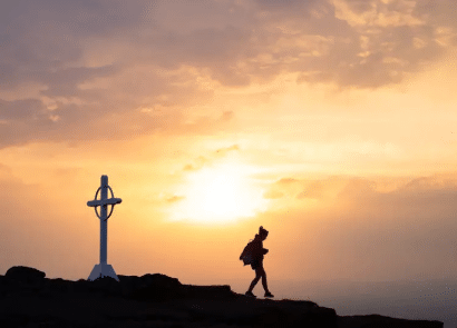 A person walks near a white Celtic cross on a rocky hilltop at sunset, with the sky glowing in warm orange and yellow hues.