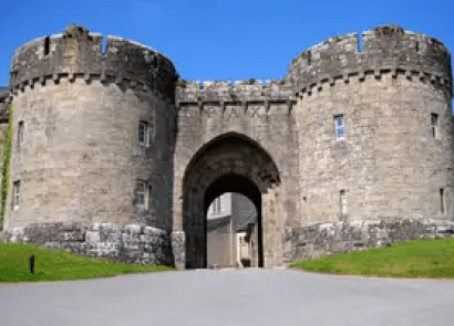 Stone castle gatehouse with two round towers and an arched entrance, set against a clear blue sky, with grass and a paved road in the foreground.