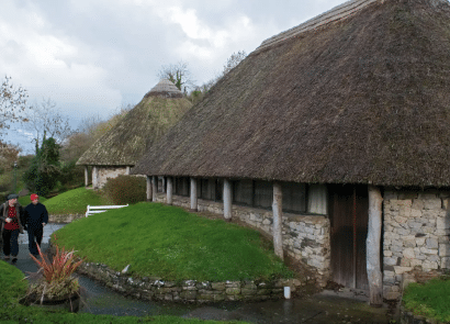 Two people walk on a path beside a stone cottage with a thatched roof, surrounded by green grass and trees. Another similar cottage is visible in the background under a cloudy sky.