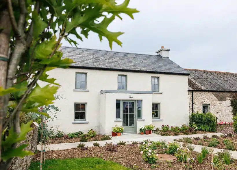 A two-storey white cottage with grey trim and a slate roof, surrounded by a tidy garden with plants and flowers. A tree branch is in the foreground, partially framing the house. The sky is overcast.
