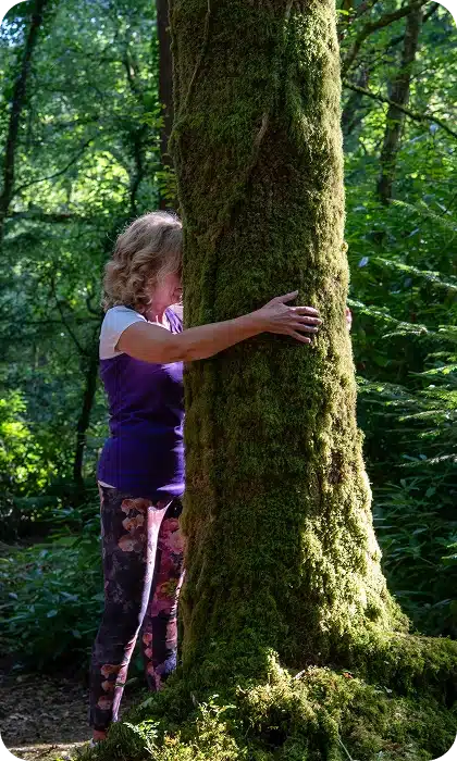 A woman with curly blonde hair wearing a purple shirt and floral leggings hugs a large, moss-covered tree in a sunlit forest. Lush green foliage surrounds her in the background.