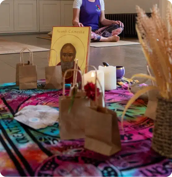 A colourful cloth with candles, gift bags, and a religious icon is arranged on the floor, whilst a person sits cross-legged in the background on a yoga mat in a calm, indoor setting.