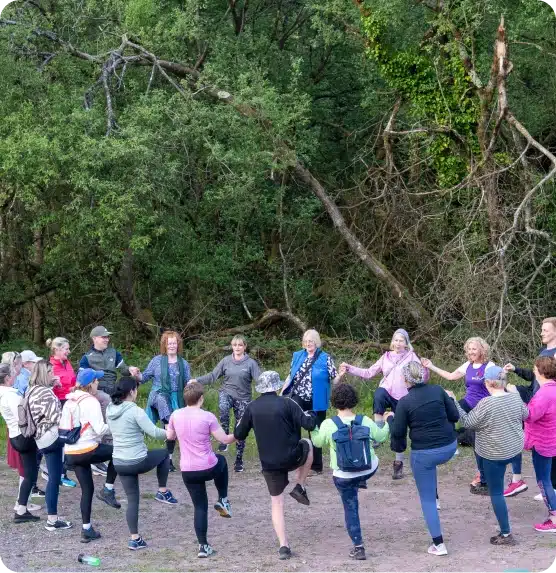 A group of people stand in a circle outdoors, holding hands and lifting one leg, surrounded by lush green trees and foliage. The group appears to be participating in a group activity or exercise in nature.
