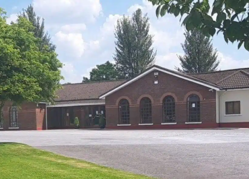 A single-storey brick building with arched windows stands beside a tree-lined drive under a sunny sky with clouds. Green grass and leafy trees frame the foreground.