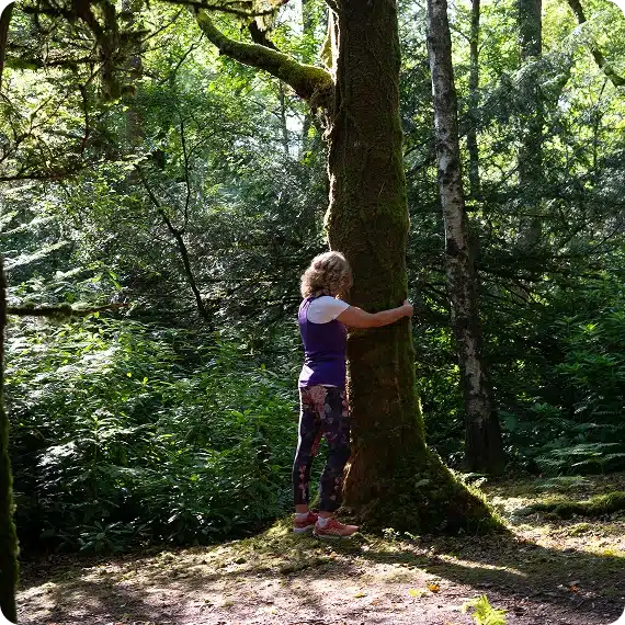 A person with curly hair, wearing a purple top and patterned leggings, hugs a mossy tree in a sunlit forest surrounded by lush green foliage.