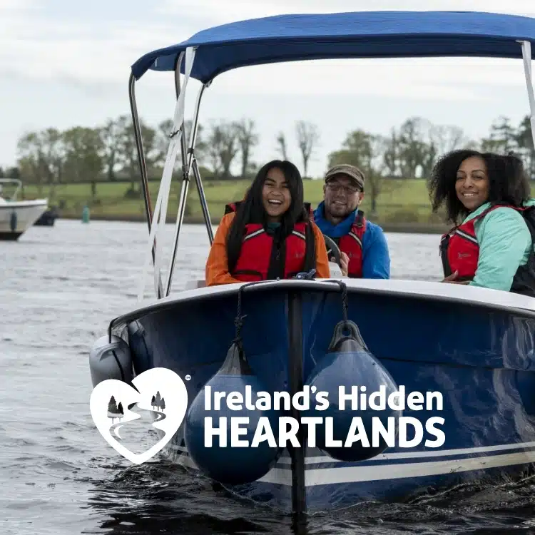 Three people wearing life jackets smile whilst sitting on a small boat on a river, with trees and another boat in the background. Text reads: "Ireland’s Hidden Heartlands" with a heart-shaped logo.