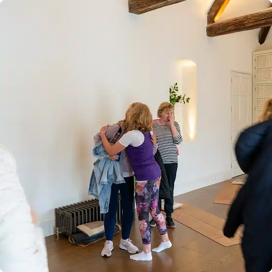 Two women hug each other in a bright room with wooden floors and exposed beams, while another woman stands nearby watching and touching her face. Yoga mats and coats are visible in the background.