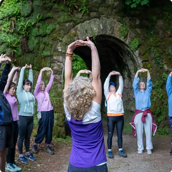 A group of people stands outdoors in front of a stone arch, stretching with arms raised above their heads. They appear to be participating in a group exercise or yoga session in a natural, green setting.