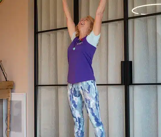 A woman with blonde hair wearing a purple top and patterned leggings stands indoors with her arms raised overhead, smiling joyfully. She is standing in front of tall glass doors with curtains.