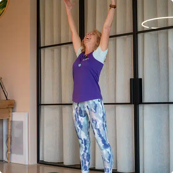 A woman with blonde hair wearing a purple top and patterned leggings stands indoors with her arms raised overhead, smiling joyfully. She is standing in front of tall glass doors with curtains.