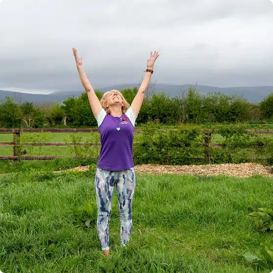 A woman standing on grass outdoors with arms raised towards the sky, wearing a purple jumper and patterned leggings, surrounded by greenery and trees with mountains and a cloudy sky in the background.