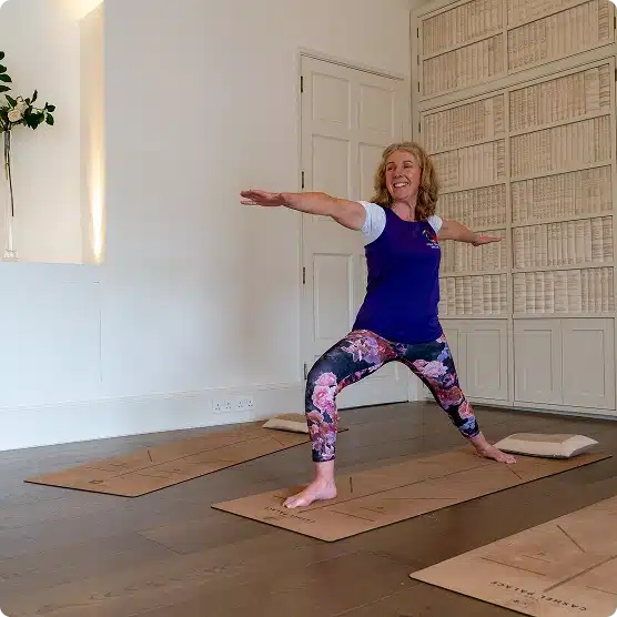 A woman practises yoga in a bright room, standing on a mat in the Warrior II pose, smiling, with arms extended. The room has wooden floors, white walls, and shelves filled with books.