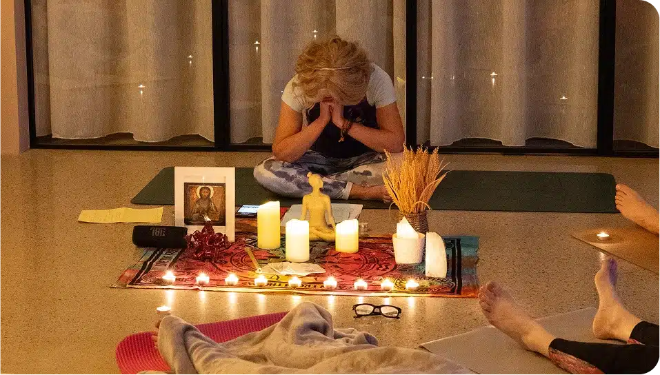 A person sits cross-legged, head bowed and hands in prayer, in front of a small altar with candles, a statue, framed photo, and dried plants. Another person's legs are visible in the foreground, suggesting a group setting.