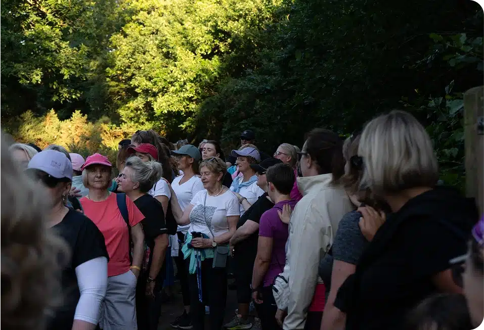 A group of women, some wearing hats and casual clothing, stand closely together outdoors on a wooded path, surrounded by green trees and shaded areas. The mood appears relaxed and sociable.