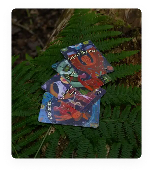 A small stack of colourful oracle cards lies on bright green ferns, resting on a mossy log. The top card reads "Expect the Best" and others display words like "Meditate" and "Call on Joy.