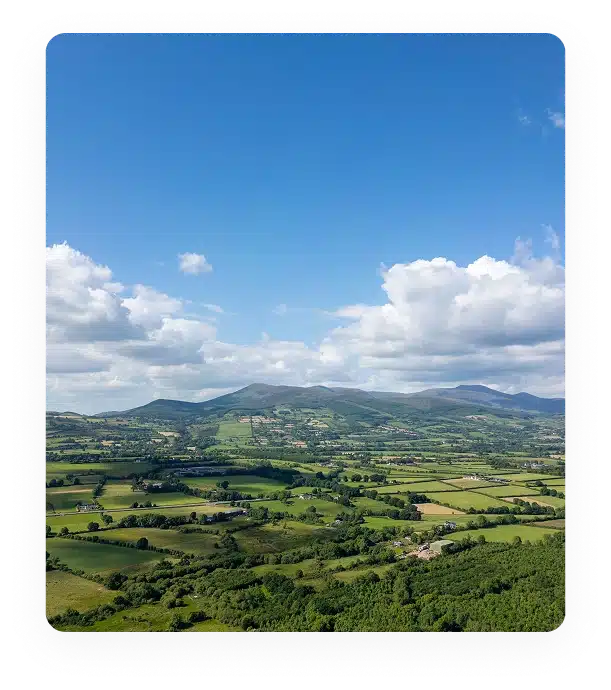 A scenic aerial view of green fields, scattered trees, and small farms with rolling hills and mountains in the background under a bright blue sky with fluffy white clouds.