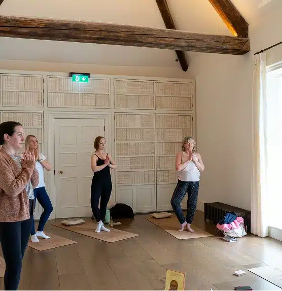 Four women stand on yoga mats in a bright room with wooden floors and exposed beams, practising yoga poses together. Shelves of books line the wall behind them. Natural light comes in through large windows.