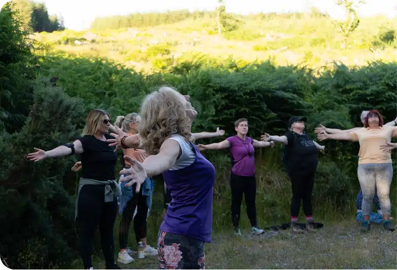 A group of people stand outdoors in a circle with arms outstretched, participating in a nature or relaxation activity surrounded by greenery and trees.