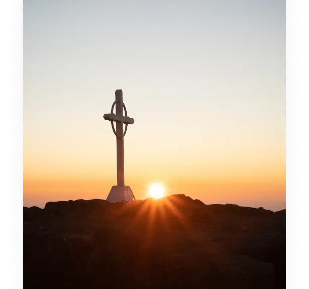A metal cross stands on rocky ground at the peak of a hill or mountain, with the sun setting or rising on the horizon, casting an orange glow and rays across the sky.