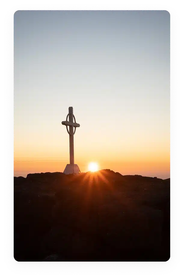 A metal cross stands on rocky ground at the peak of a hill or mountain, with the sun setting or rising on the horizon, casting an orange glow and rays across the sky.