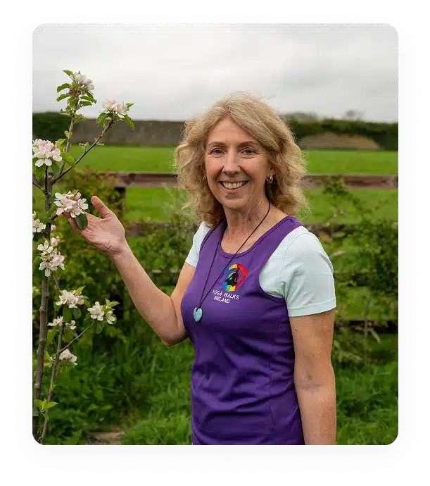 A smiling woman with curly blonde hair stands outdoors by a blossoming tree branch, wearing a purple waistcoat over a light blue shirt. Green fields and a wooden fence are visible in the background.
