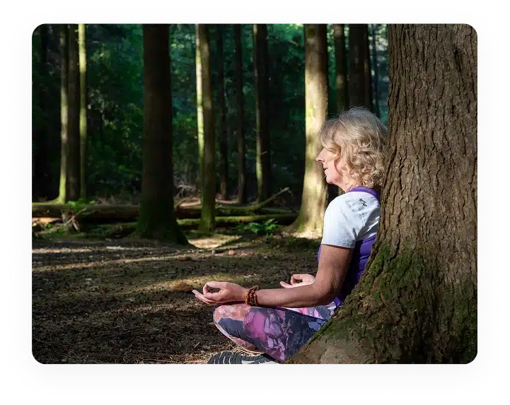An older woman with blonde hair sits cross-legged against a tree in a sunlit forest, meditating with her eyes closed and hands resting on her knees in a peaceful pose.
