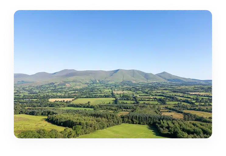 A panoramic view of patchwork green fields and clusters of trees with rolling hills and mountains in the distance, under a clear blue sky.