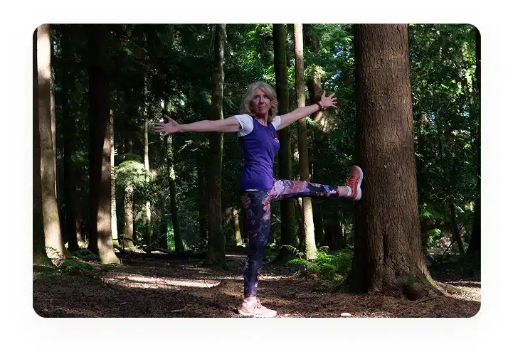 A woman stands in a forest, stretching with one leg raised and arms outstretched to the sides. She is wearing a purple shirt, patterned leggings, and pink trainers. Sunlight filters through the trees.