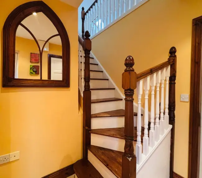 A wooden staircase with white balusters and dark handrails leads to an upper floor in a house with yellow walls, chequered tiled floor, and an arched mirror hanging on the wall.