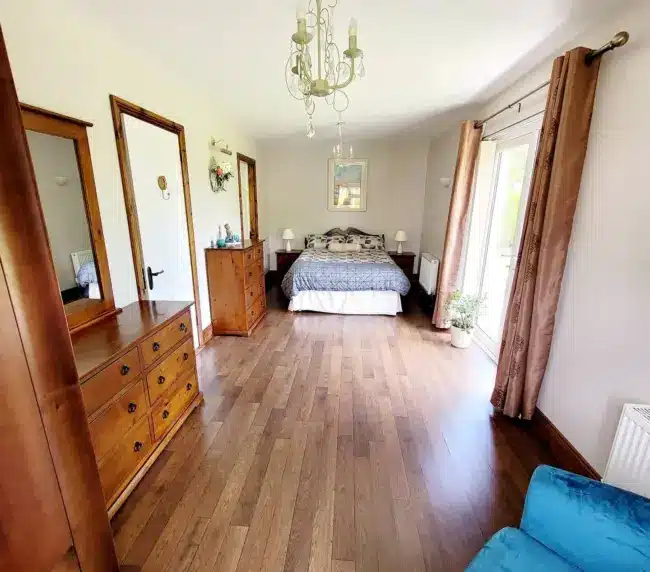 Bright bedroom with wooden floors, brown furniture, a bed with patterned bed linen, mirrored wardrobes, a blue armchair, large window with brown curtains, and a chandelier hanging from the white ceiling.