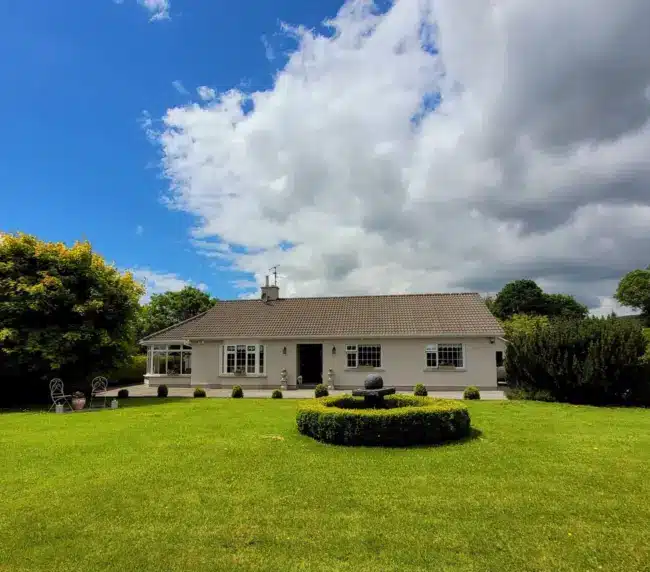 A single-storey white house with a grey roof sits behind a large, well-manicured green lawn. There is a circular garden feature in the foreground, trees on each side, and a partly cloudy blue sky overhead.
