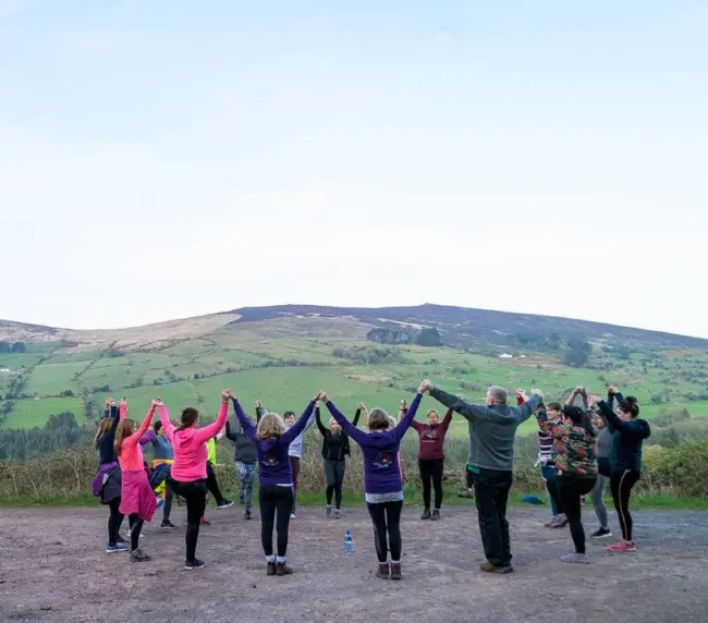 A group of people stand outdoors in a circle, holding hands with arms raised, facing green hills and a blue sky in the background. They appear to be enjoying a group activity in nature.