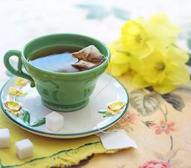 A green teacup with tea and a teabag sits on a matching saucer decorated with yellow flowers. Three sugar lumps and a yellow serviette are nearby, with yellow daffodils in the background on a floral tablecloth.